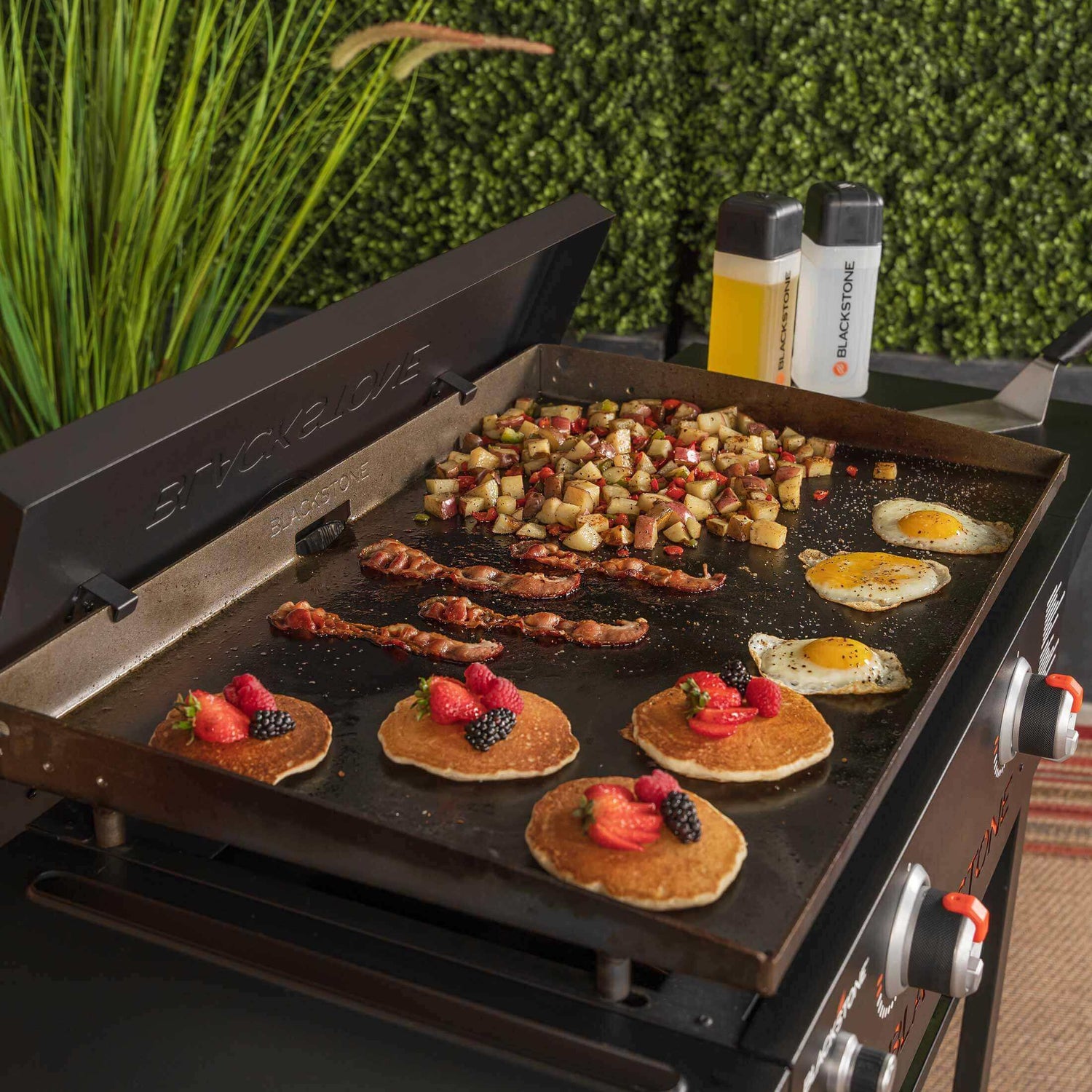 Blackstone 28” ParilaGrilli cooking breakfast foods including pancakes, eggs, bacon, and potatoes with greenery in the background