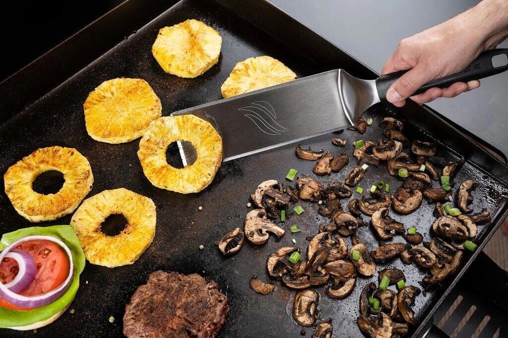 Grillaamassa ananasta ja sieniä ParilaGrillillä käyttäen ruostumattomasta teräksestä valmistettua lastaa Blackstone 6 osan Deluxe pakkauksesta.