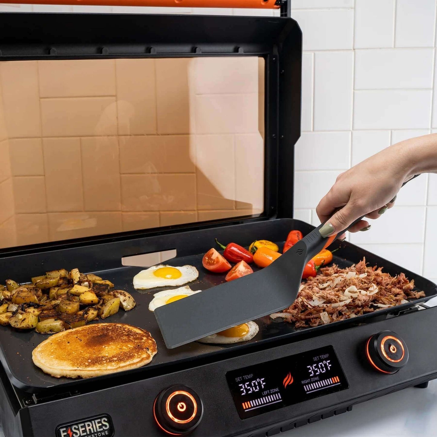 Aamiaisen valmistaminen Blackstone E-Series -grillillä käyttäen Blackstone 4-osaista työkalusettiä, johon kuuluu lasta; valmistumassa pannukakkuja, munia ja vihanneksia.