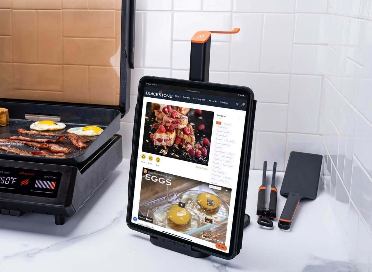 Blackstone E-Series tool holder with inserted tablet showing recipe, tools neatly arranged on kitchen counter near cooking griddle.