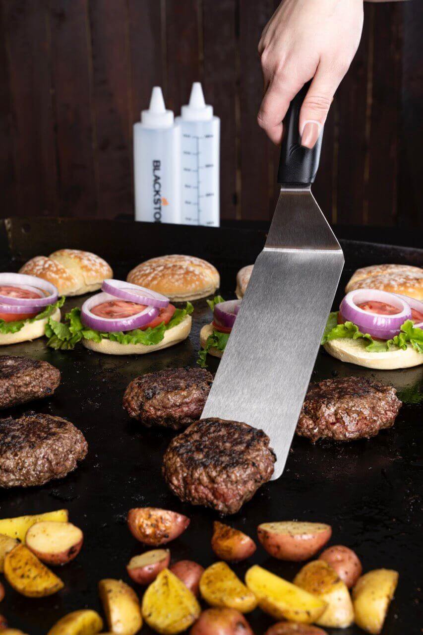 Grillaamassa hampurilaisia ja vihanneksia Blackstone ParilaGrilli -aloituspaketilla, joka sisältää ruostumattomasta teräksestä valmistetun lastan ja pullot.