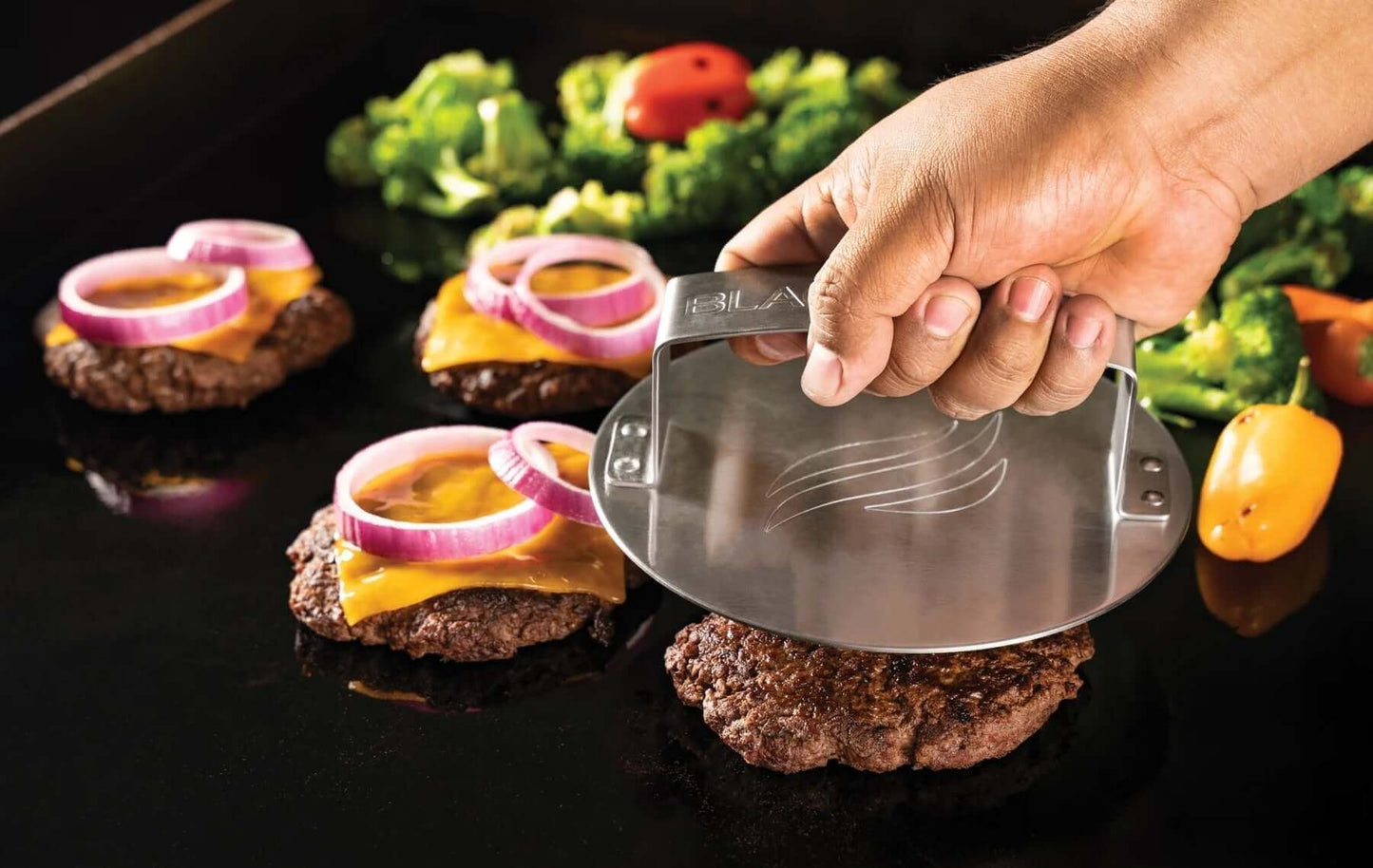 Blackstone Hampurilaispakkaus puristamassa hampurilaispihviä grillillä, taustalla vihanneksia